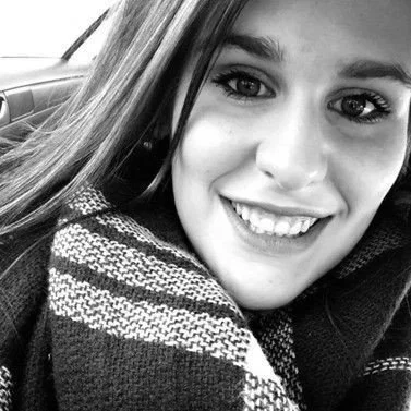 Close-up photo of a smiling young woman with long hair, wearing a patterned scarf, sitting inside a car.