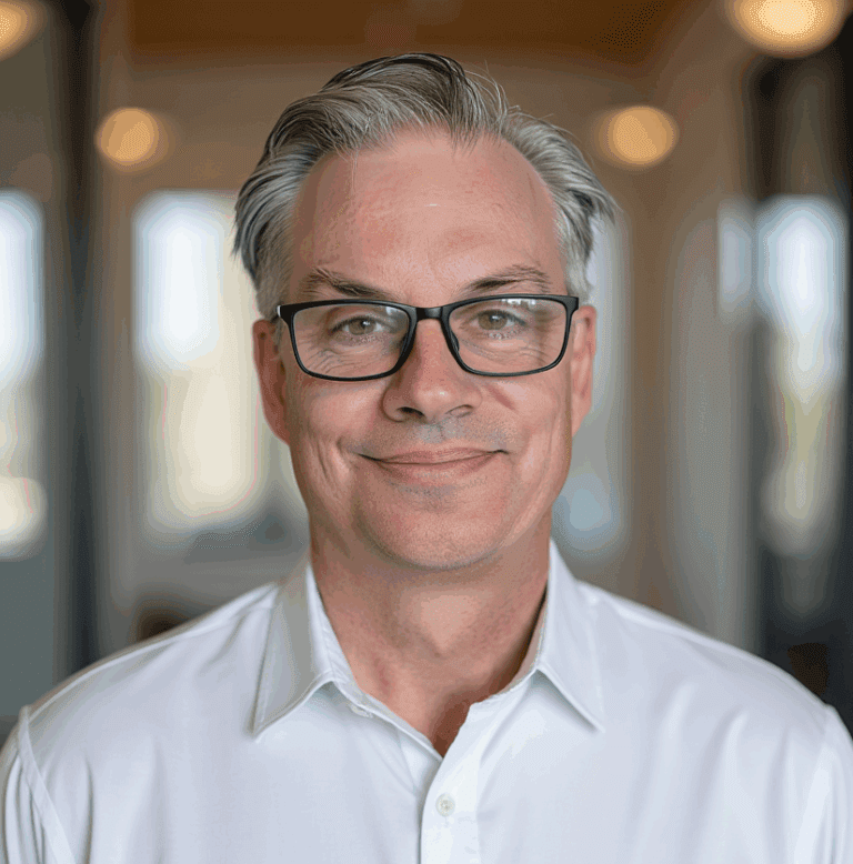 A middle-aged man with gray hair, glasses, and a white dress shirt smiling in a well-lit indoor setting.
