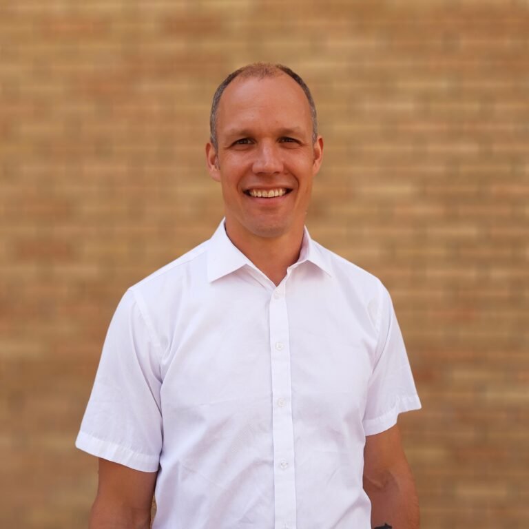 A smiling man in a white button-up shirt standing in front of a brick wall.