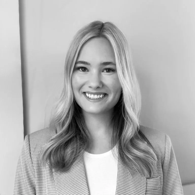 A smiling woman with long blonde hair in a checkered blazer and a white top, standing in front of a plain wall.