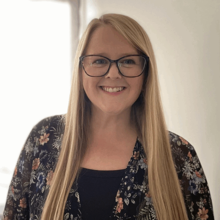 A woman with long blonde hair, wearing glasses and a floral patterned top, smiling in an indoor setting.