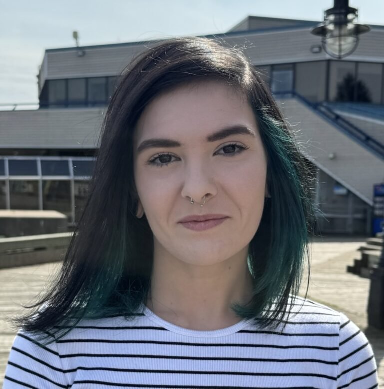 Young woman with dark hair and teal highlights, wearing a striped shirt, standing outdoors in front of a modern building.