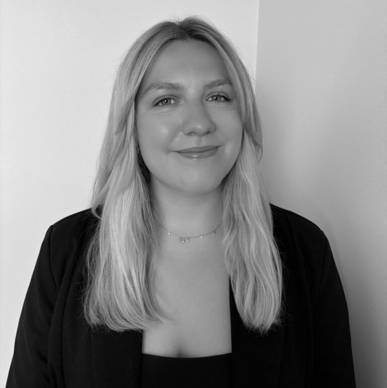 A young woman with long blonde hair, smiling, wearing a black blazer and a delicate necklace, standing against a plain wall.