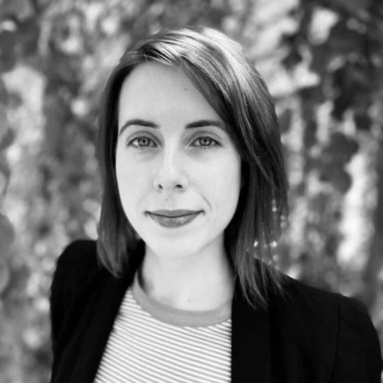 Black and white portrait of a woman with shoulder-length hair, wearing a striped shirt and blazer, smiling slightly, with a blurred background of trees.
