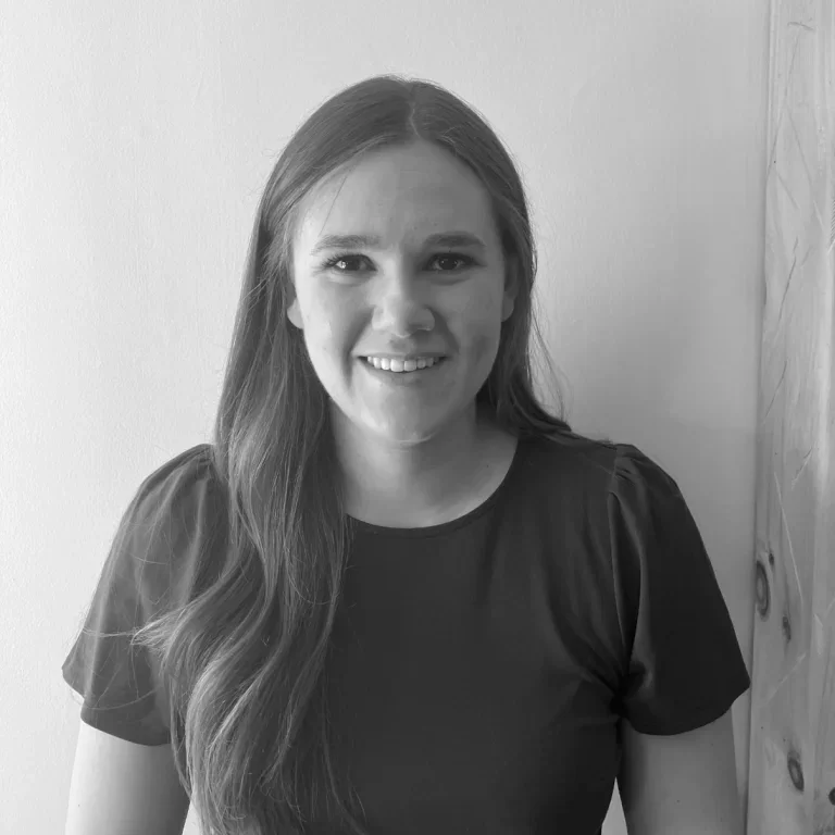 A woman with long hair smiling, wearing a dark shirt, standing against a plain wall.