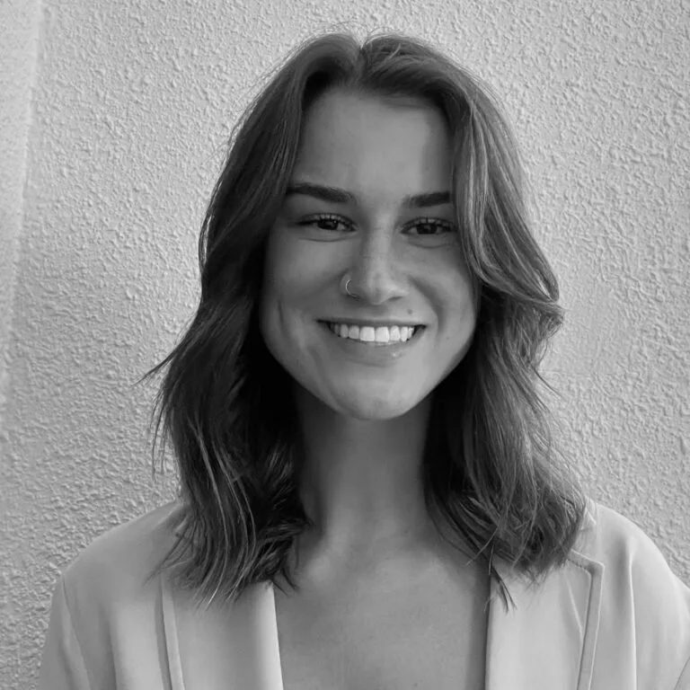A young woman with wavy hair, smiling, and wearing a nose ring, standing against a textured wall.