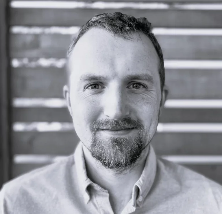 Black and white portrait of a man with short hair, beard, and mustache, wearing a collared shirt, smiling, with wooden slats background.