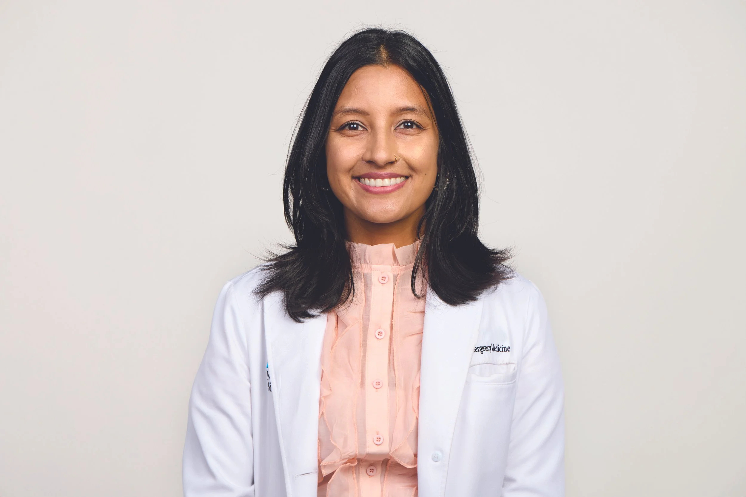 Portrait of a woman with shoulder-length black hair, smiling, wearing a white medical coat over a pink shirt, standing against a plain light gray background.