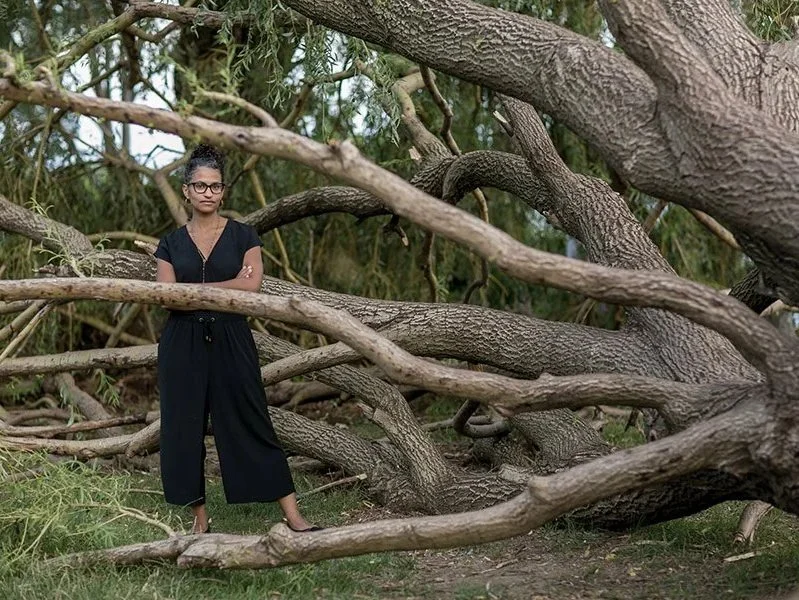 A woman with glasses and black attire standing amidst large tree branches in a natural outdoor setting.