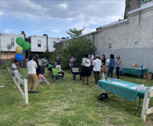 People gathered in a backyard for an outdoor event, with some seated and others standing, and colorful balloons tied to a white fence.