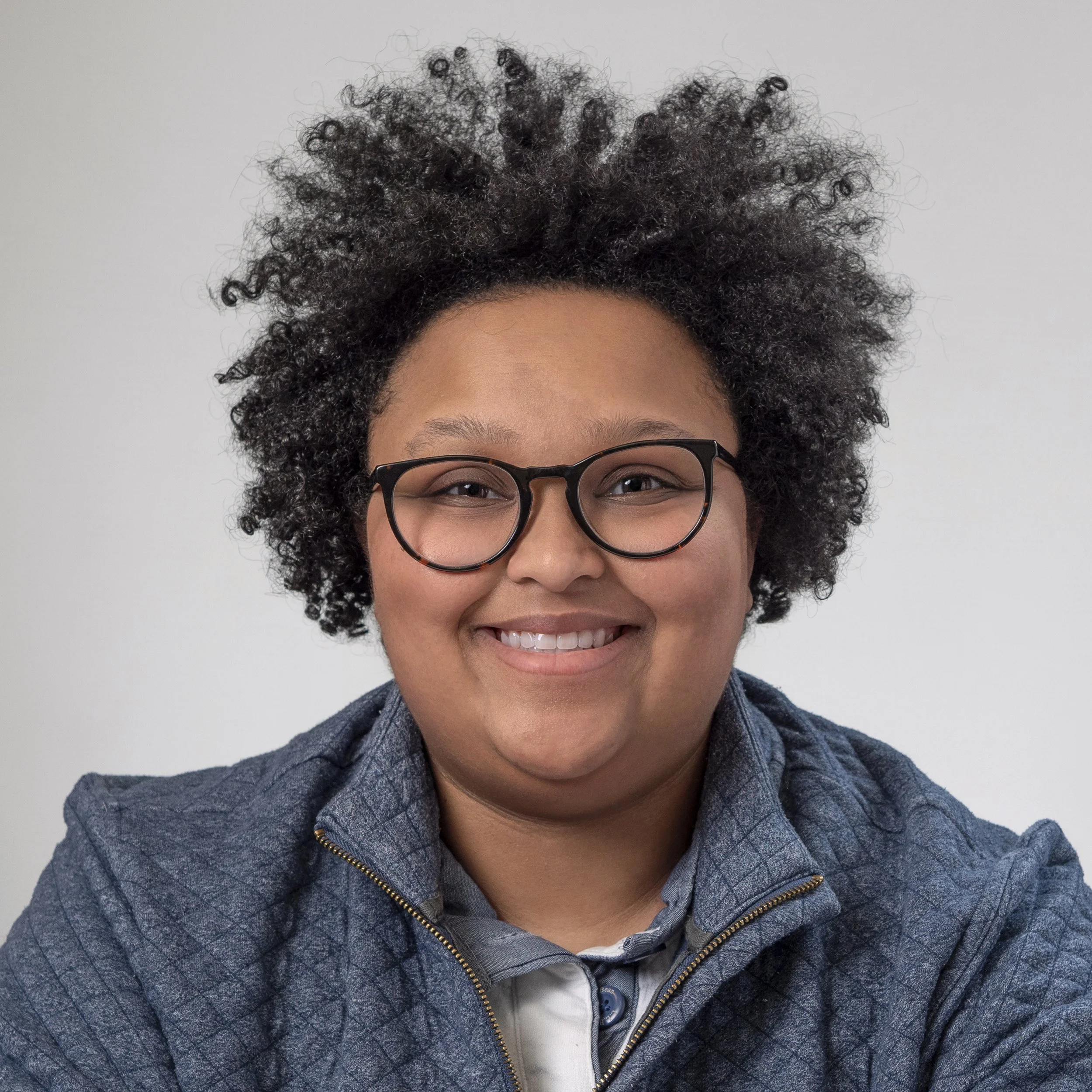 Close-up of a person with curly hair, wearing glasses and a gray patterned jacket, smiling.