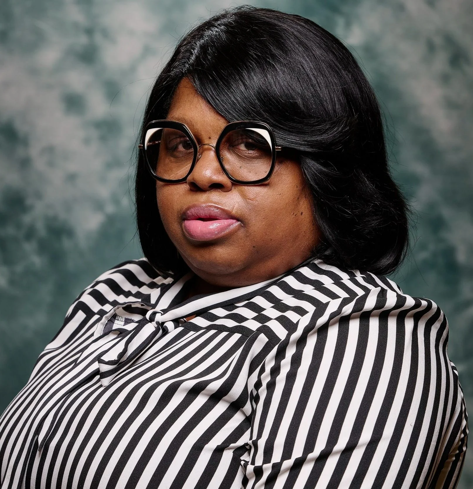 A woman with black hair styled in a bob is wearing large black glasses and a black and white striped shirt, sticking her tongue out slightly, with a grayish green textured background.