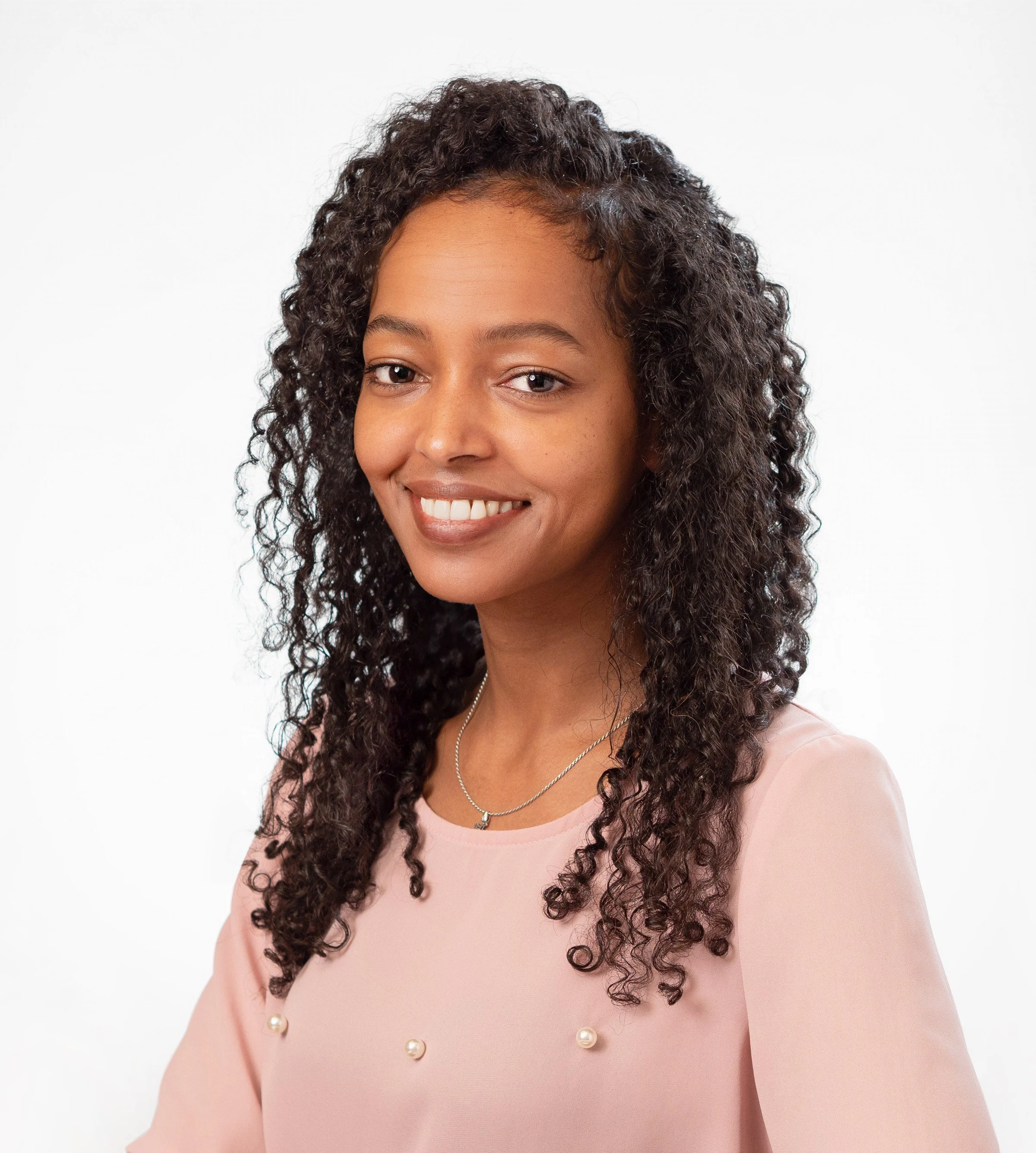Close-up of a young woman with curly brown hair, smiling softly, wearing a light pink blouse with pearl embellishments, and a silver necklace, against a plain white background.