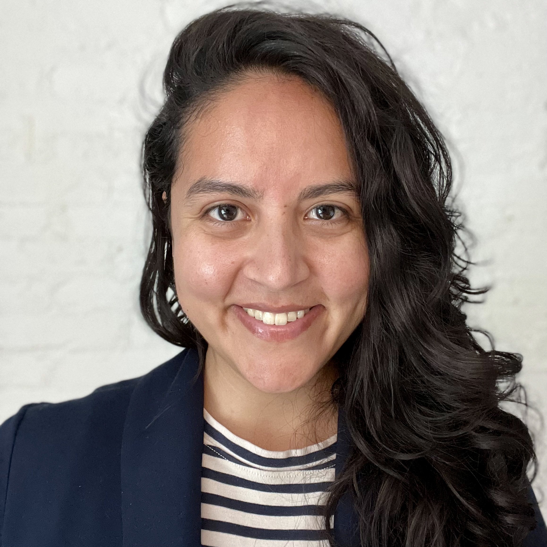 Close-up of a smiling woman with long, dark hair, wearing a navy blazer over a striped shirt, standing against a light-colored background.