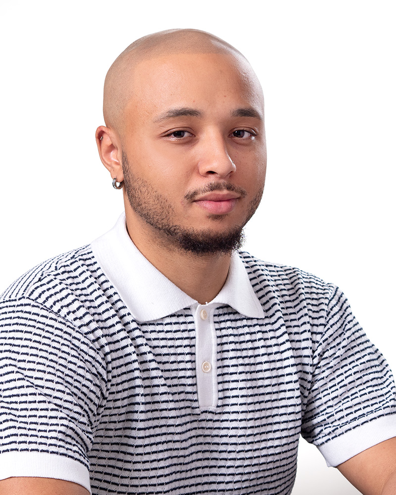 Portrait of a young man with a shaved head and beard wearing a white and black striped polo shirt, looking at the camera with a slight smile.