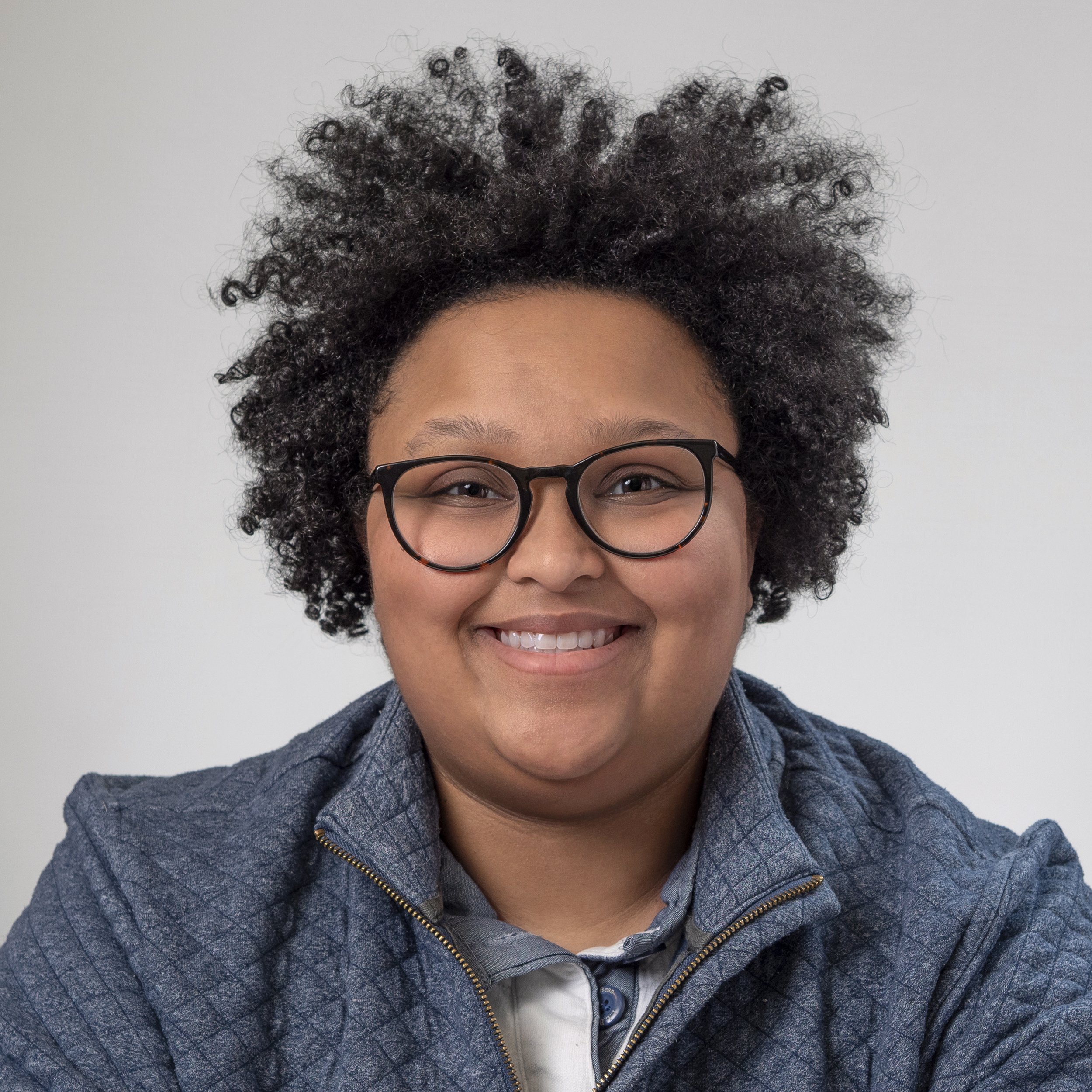 Close-up of a person with medium skin tone wearing glasses and a denim jacket, smiling, with curly hair and a gray background.
