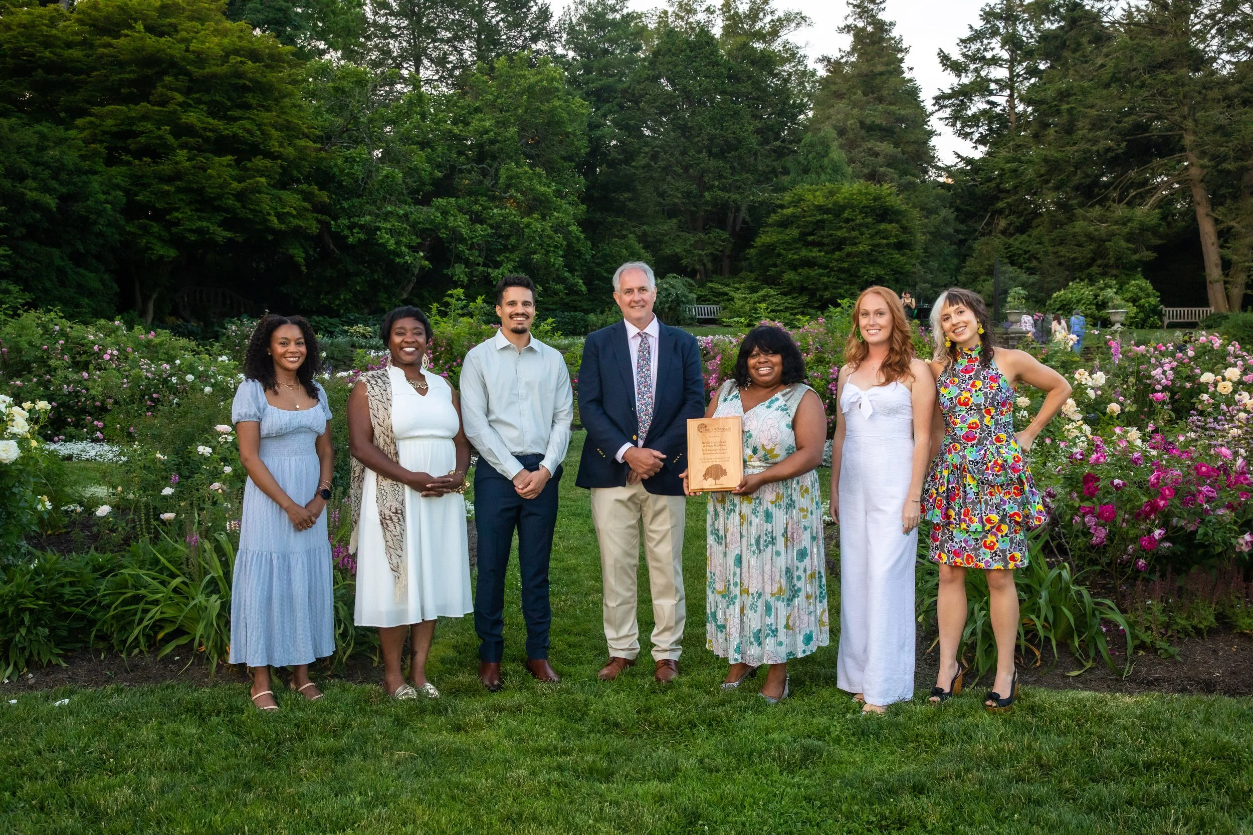A group of eight diverse people: six women and two men, standing together outside in a garden with flowers and tall trees. They are smiling and dressed in summer clothing, and one person is holding a framed award.