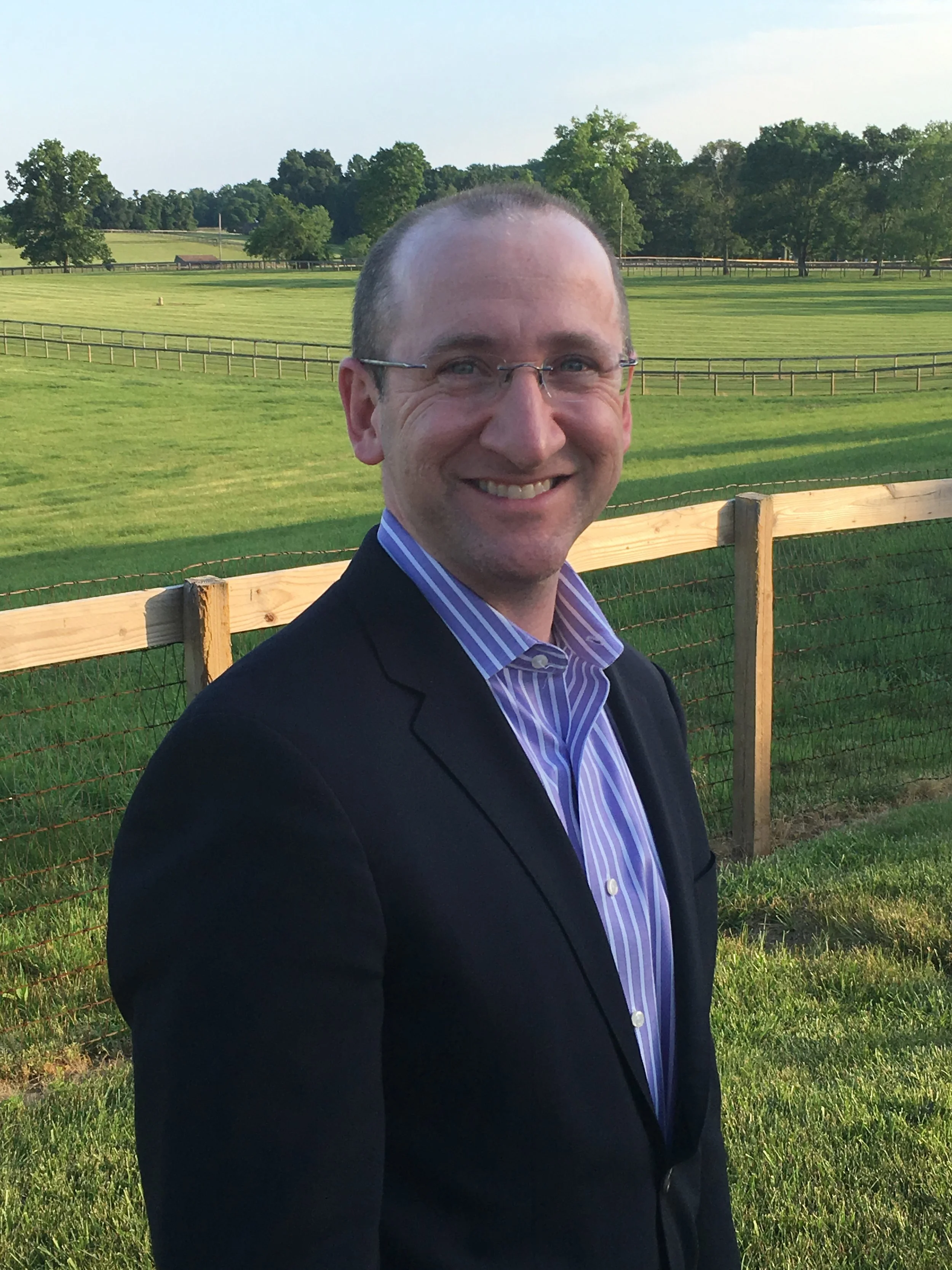 A smiling man with glasses wearing a dark blazer and a purple striped shirt outdoors, with a green field, trees, and a fence in the background during daytime.