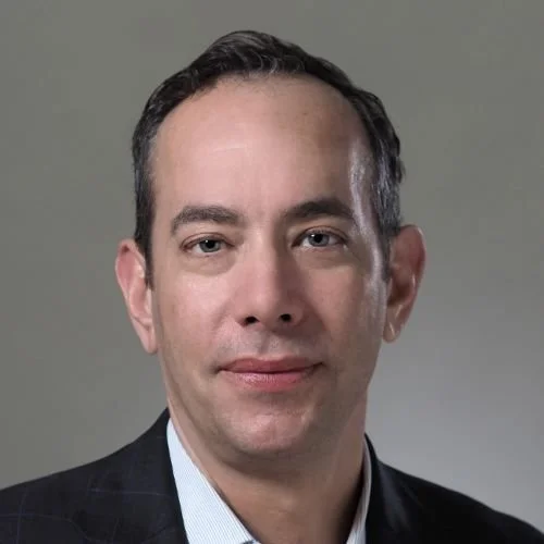 Headshot of a man with short dark hair, wearing a dark suit and light blue shirt, against a neutral background.