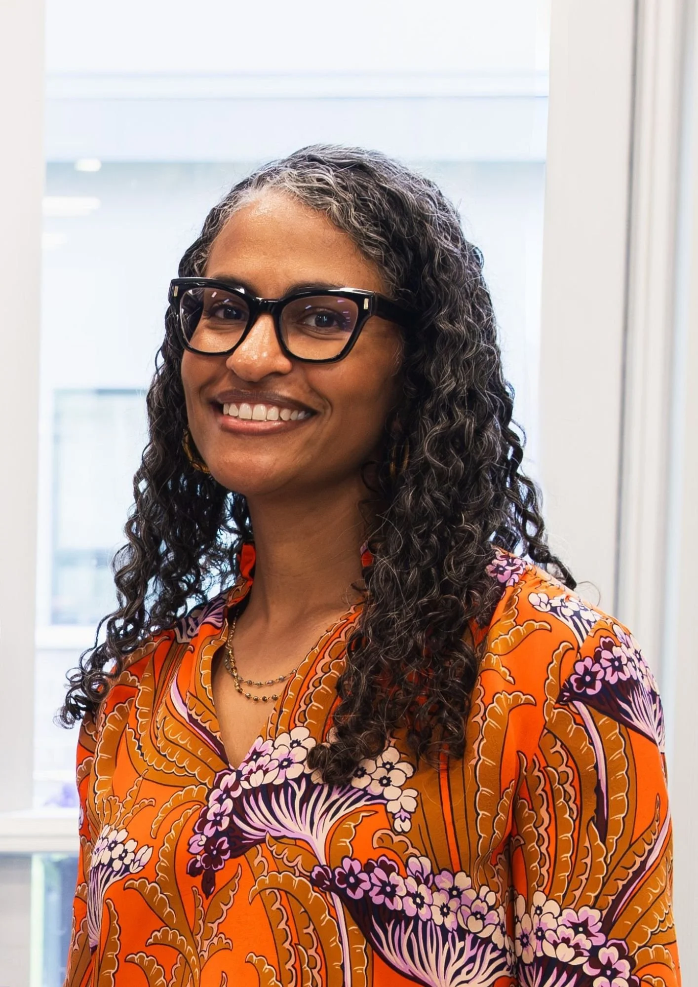 A woman with long curly hair and glasses smiling in front of a window, wearing an orange floral-patterned blouse and layered necklaces.