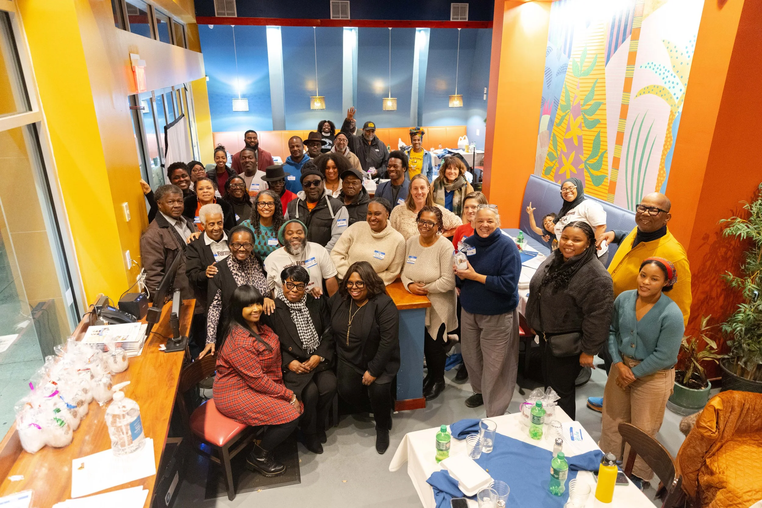 A large group of diverse people gathered in a decorated restaurant dining area, smiling and posing for a group photo. There are tables with food and drinks around them, and colorful wall art and lighting overhead.