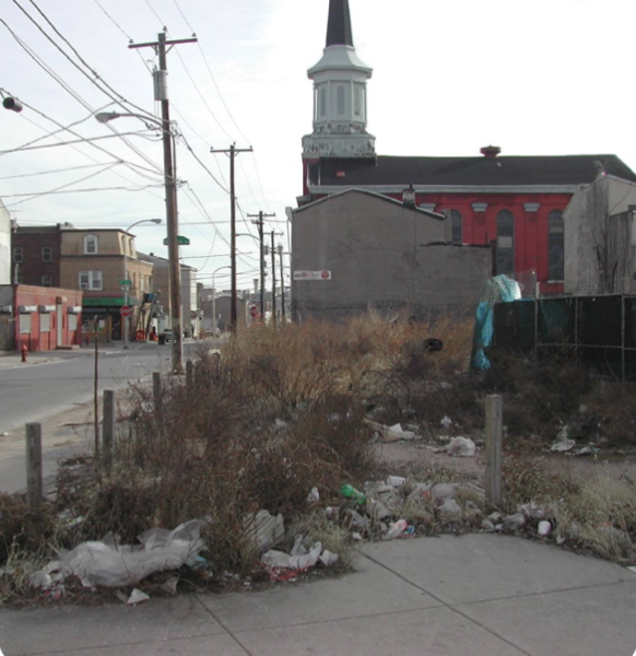 Empty lot with trash and overgrown weeds in an urban area, with power lines and a church with a steeple in the background.