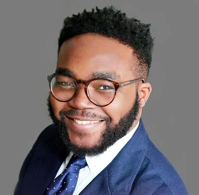 Portrait of a smiling man with glasses, a beard, and curly hair, wearing a navy suit and polka dot tie, against a gray background.