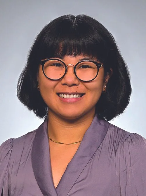 Headshot of an Asian woman with short black hair, wearing glasses, a purple blouse, and a gold necklace, smiling. 