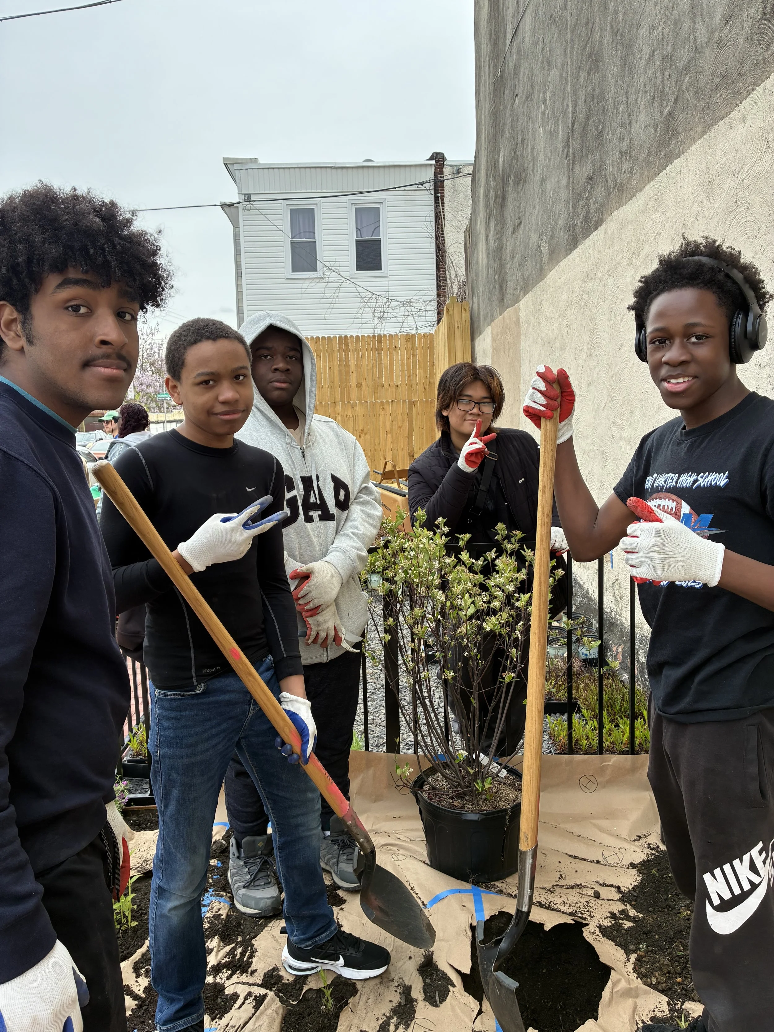 Group of five young people planting a shrub in a garden, with gardening gloves, tools, and soil visible, against a backdrop of a house and a wooden fence.