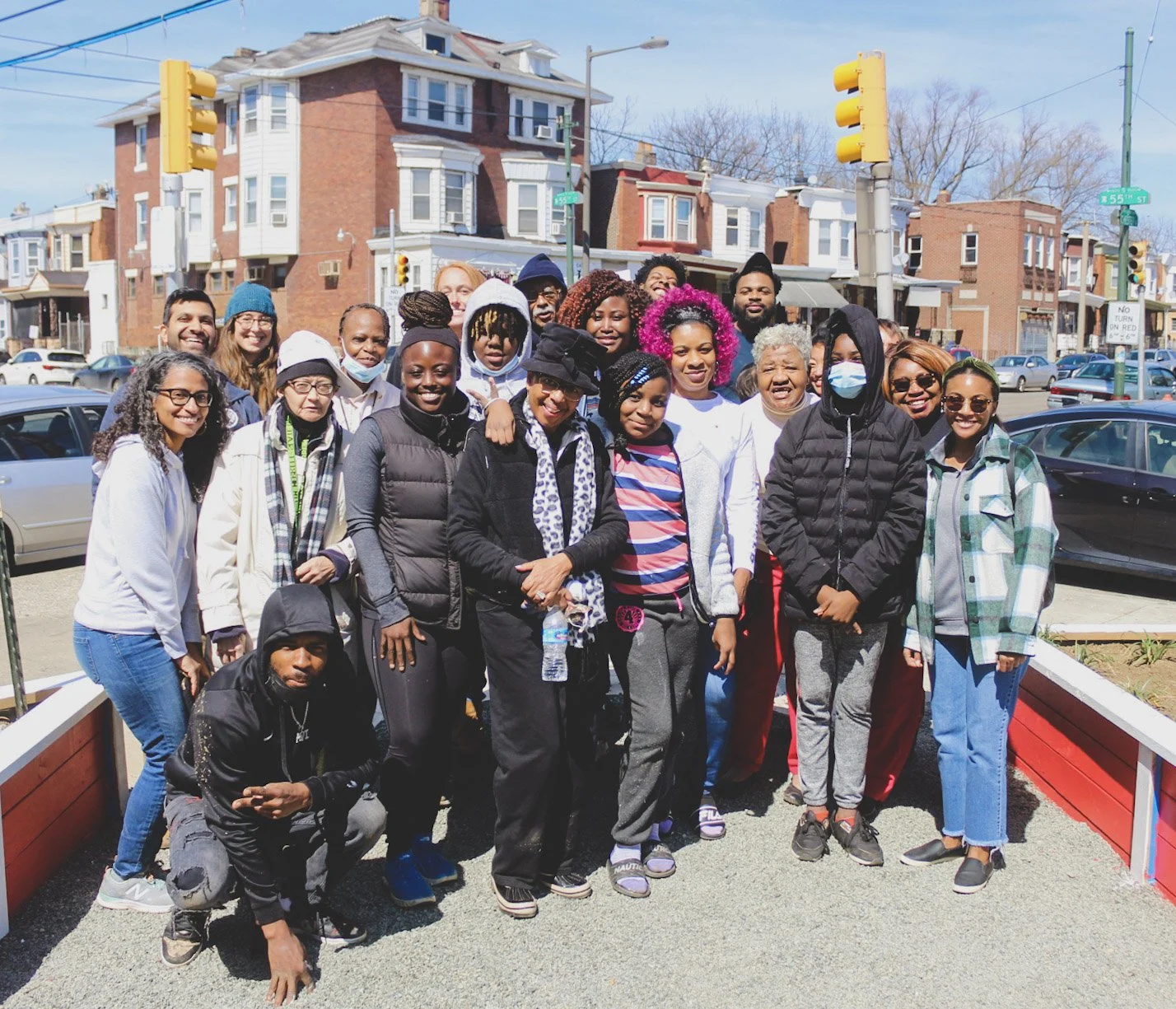 A diverse group of people, mostly women and some men, standing outdoors on a sunny day in front of a city street with buildings, cars and traffic lights, posing for a photo together.