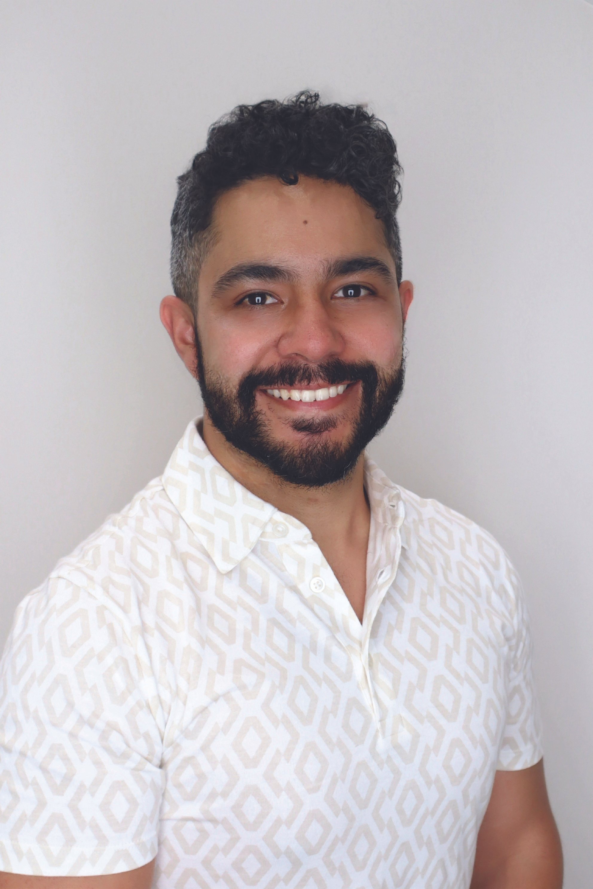 A smiling man with dark curly hair and a beard, wearing a white shirt with a geometric pattern, standing against a plain white wall.