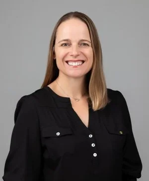 Headshot of a woman with long light brown hair, smiling, wearing a black button-up shirt, against a plain gray background.