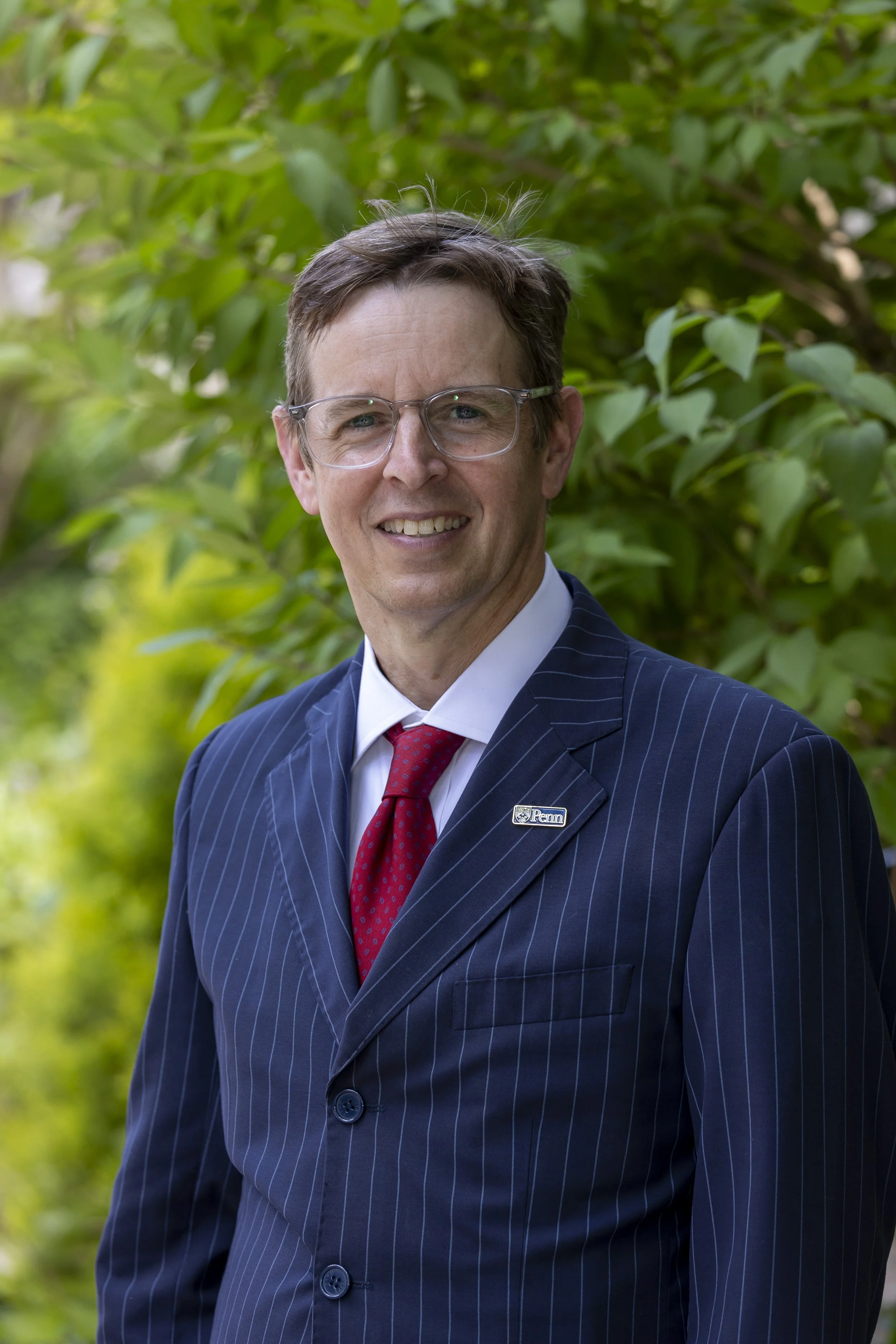 A smiling man in a pinstripe suit, white shirt, and red tie standing outdoors with greenery in the background.
