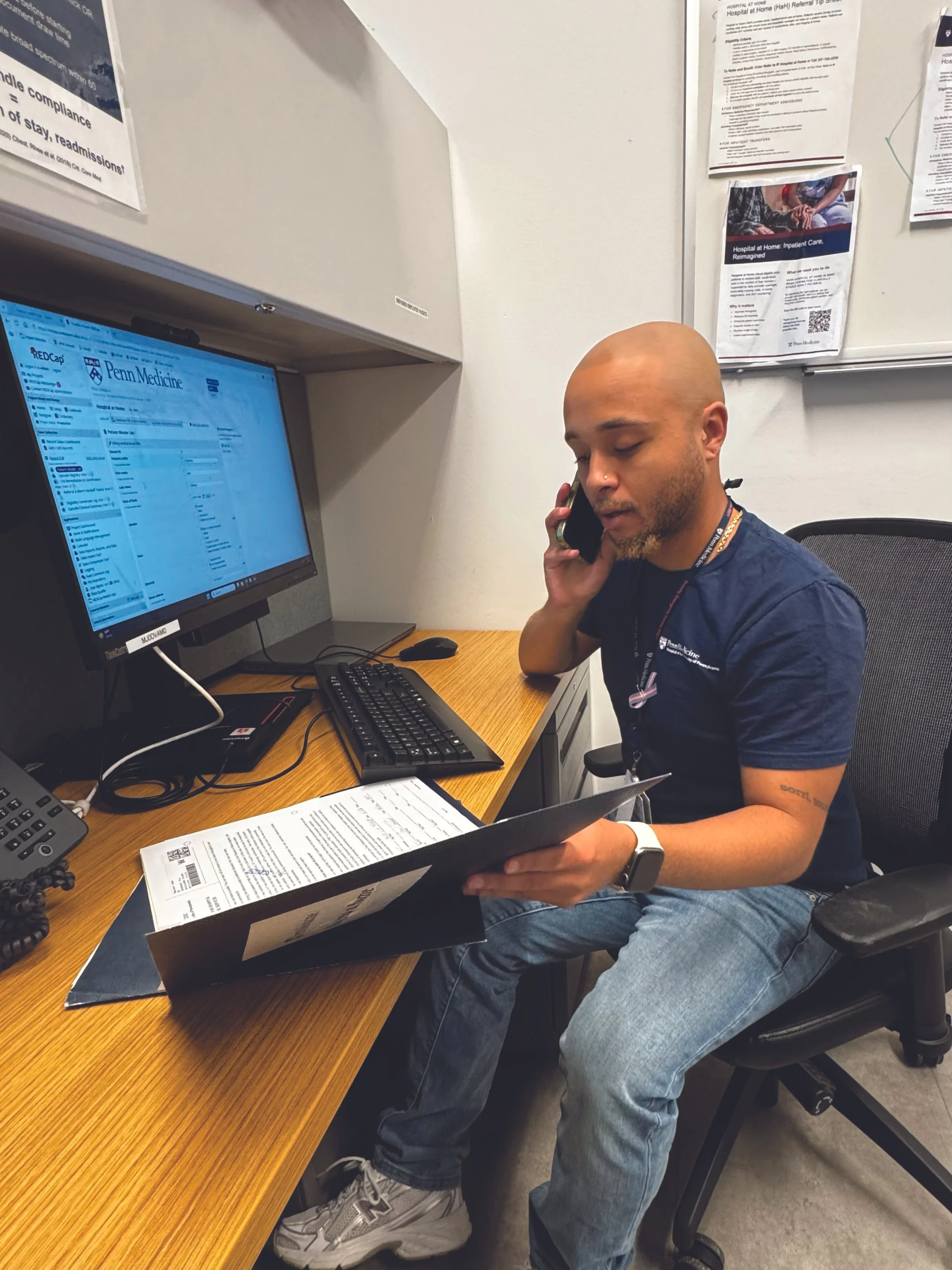 A man sitting at a desk in an office, talking on the phone while holding a pamphlet, with a computer monitor, keyboard, and papers on the desk and medical posters on the wall.