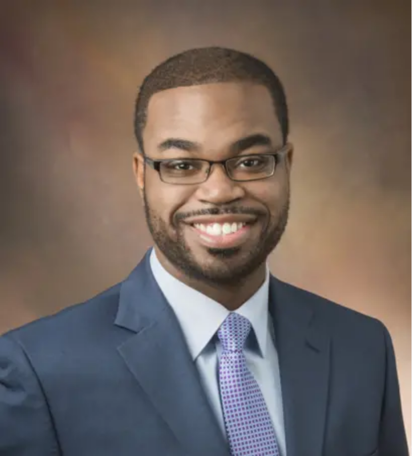 A smiling man with glasses in a blue suit, white shirt, and purple tie, posing against a brown gradient background.