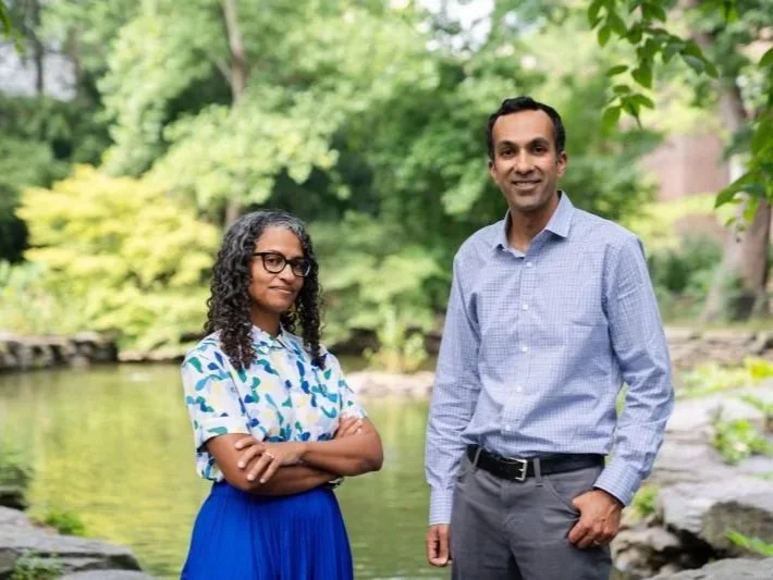 A man and a woman standing outdoors by a pond with green trees in the background.