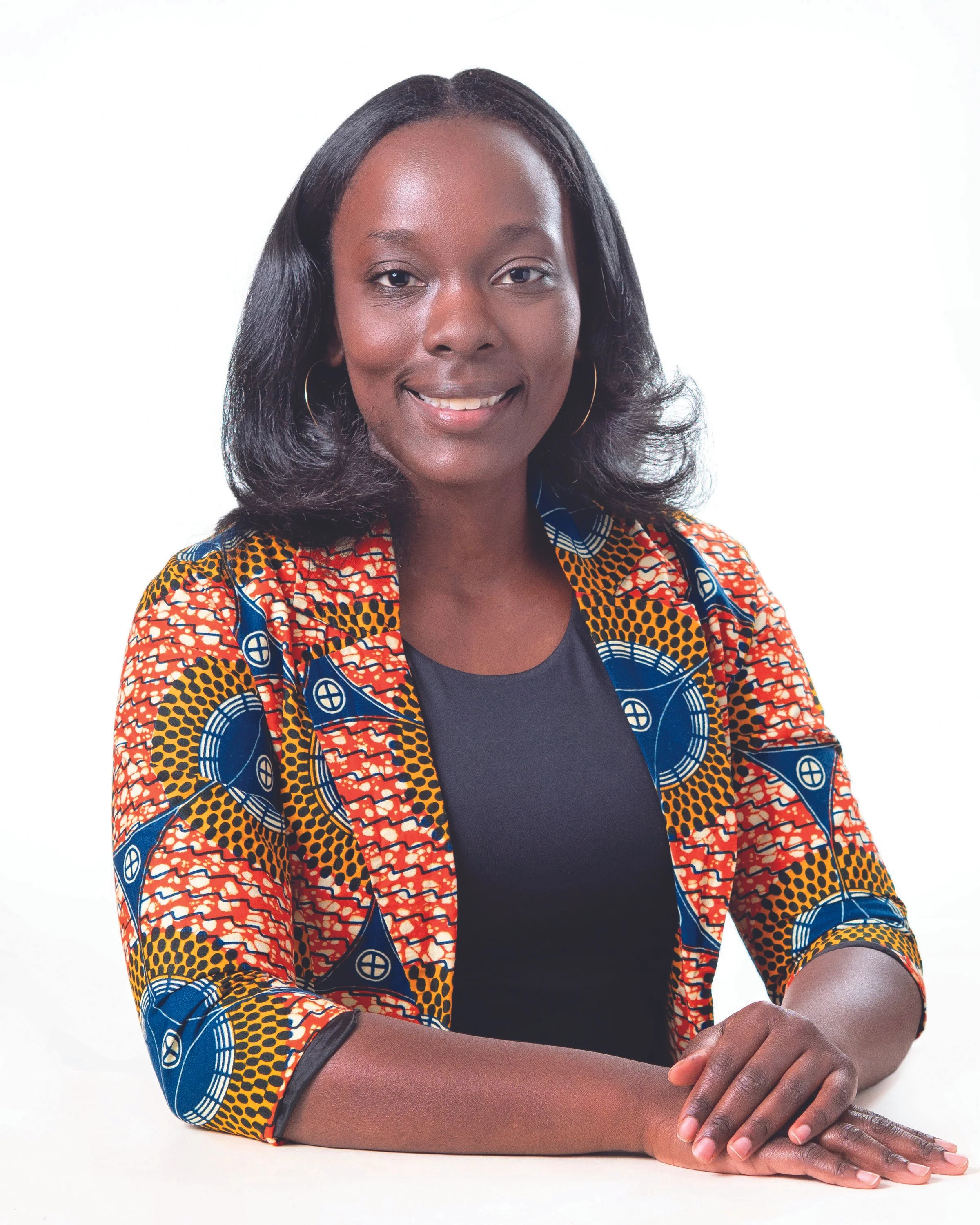 A woman with shoulder-length black hair, wearing a colorful patterned jacket with blue, orange, and yellow, smiling, sitting at a table with her hands resting on the surface, against a plain white background.