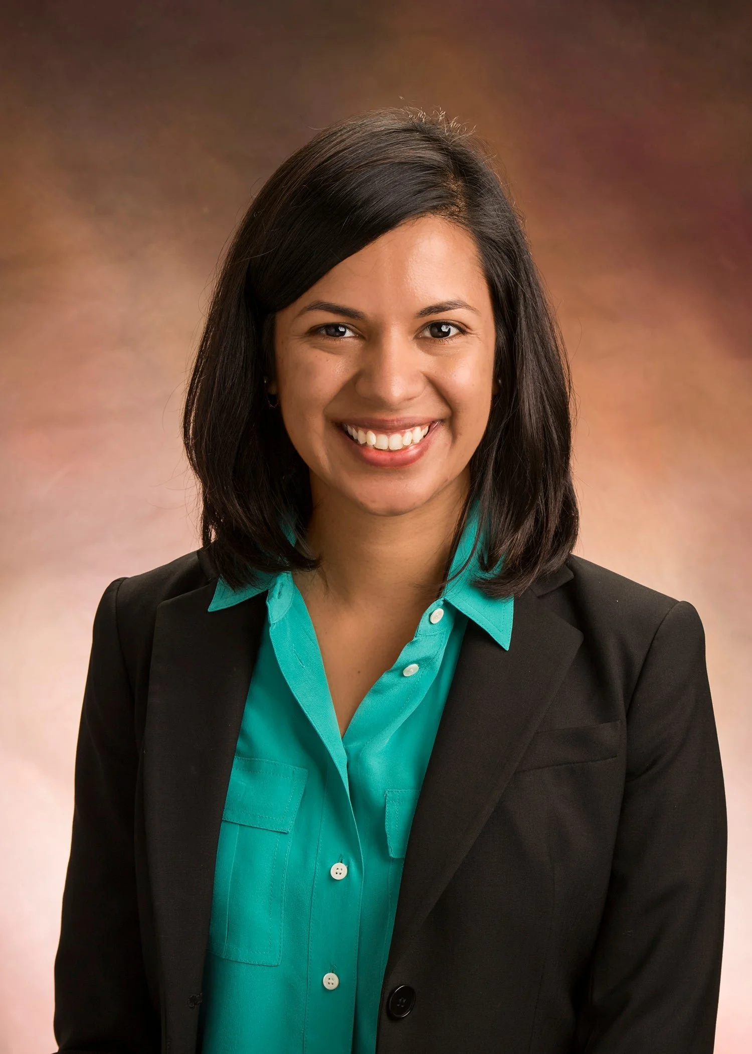 A woman with dark hair smiling, wearing a turquoise blouse and a black blazer, against a brownish gradient background.