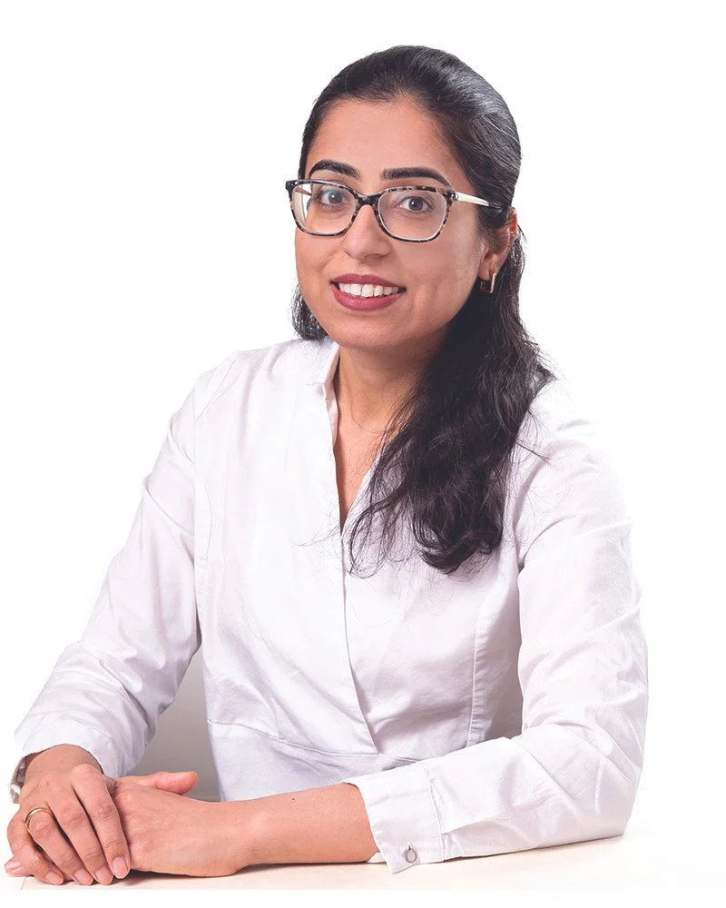 A woman with long dark hair, glasses, and a white shirt sitting at a table with hands folded, smiling at the camera against a plain white background.