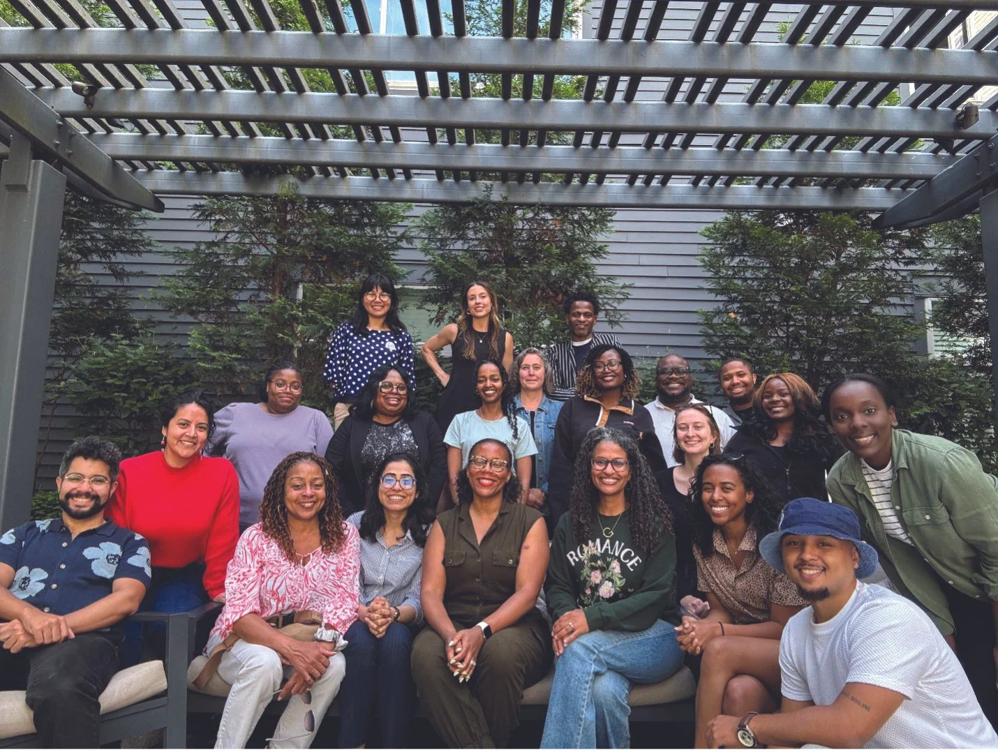 A diverse group of 20 people posing together outdoors, sitting and standing under a pergola, with trees and a gray siding wall in the background.