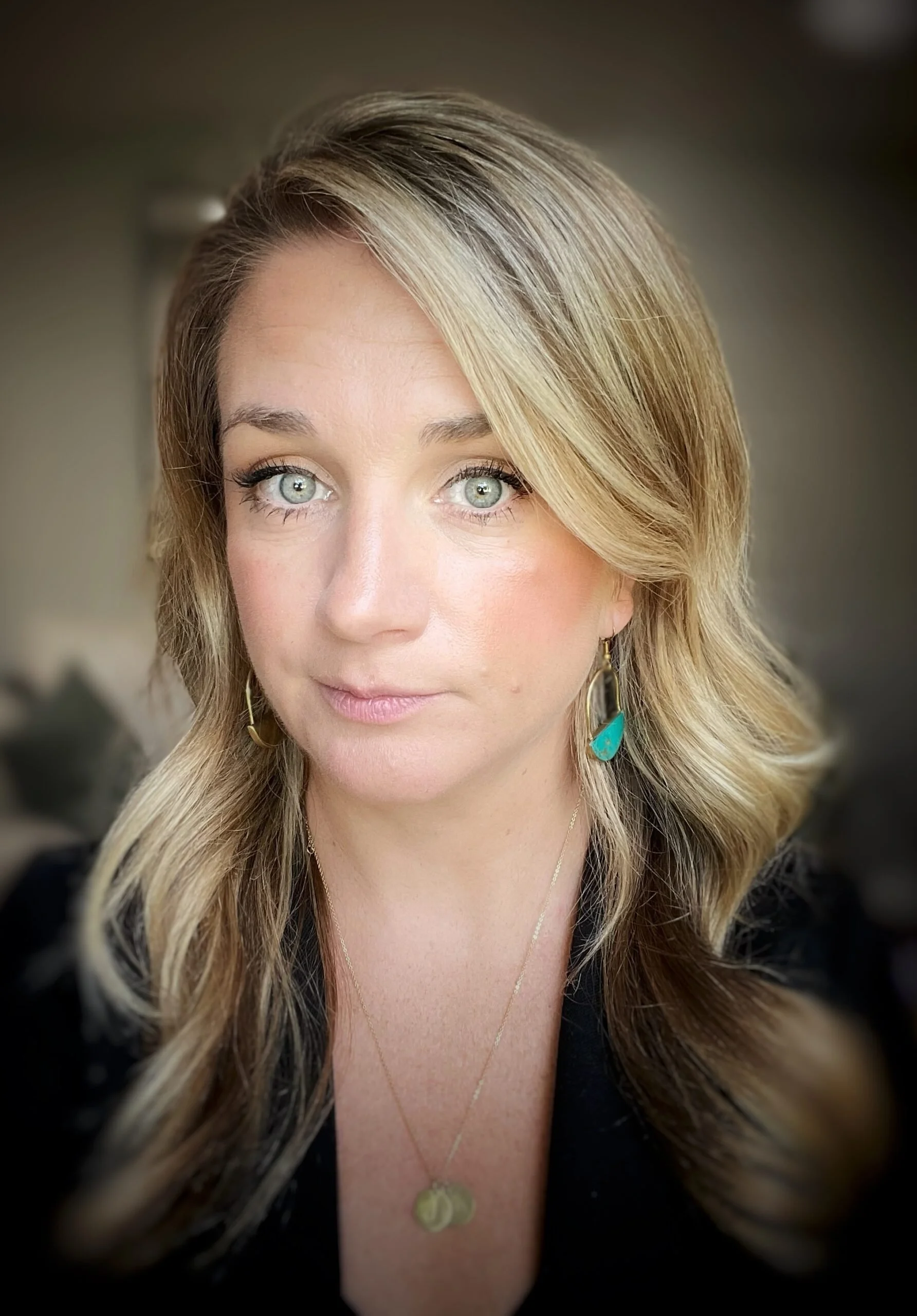 Close-up of a woman with blonde hair, blue eyes, wearing earrings and a necklace, with a black top.