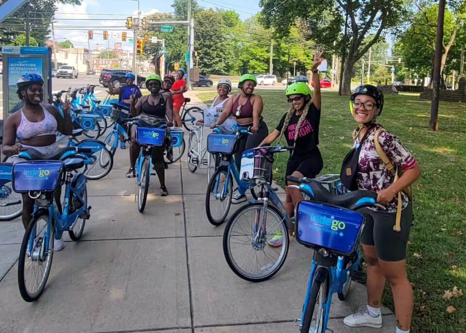 Group of smiling women with bicycles wearing helmets, standing on a sidewalk in a park, some making peace signs, with a city street and trees in the background.