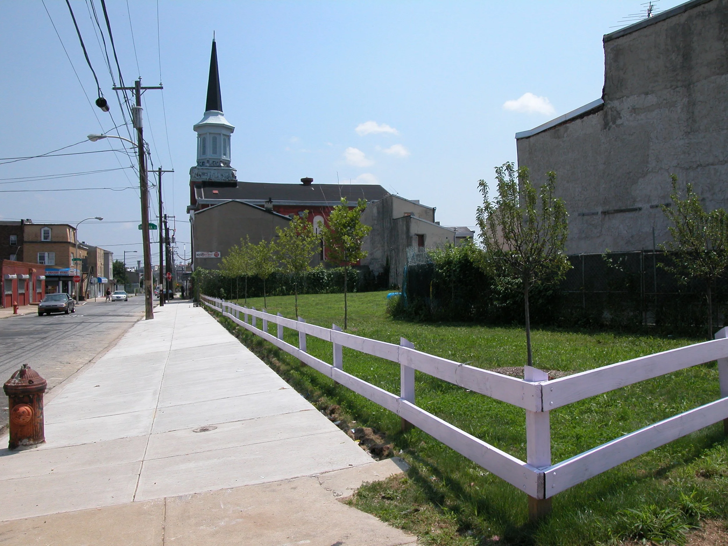 A city sidewalk with a white fence, green grass, and small trees on the right. There is a church with a tall steeple in the background, partially obscured by power lines. Several buildings and parked cars are visible along the street.