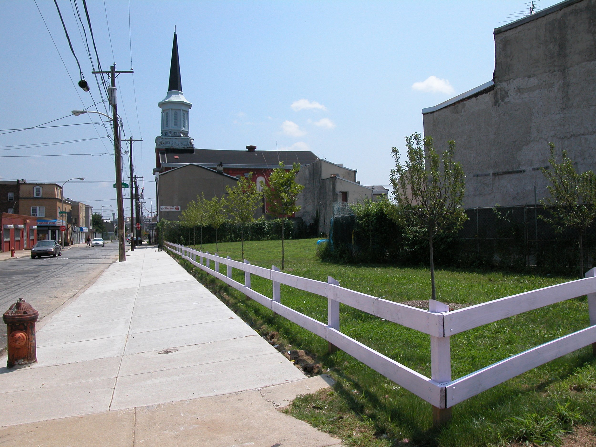 A sunny urban street with a church featuring a tall steeple in the background. The sidewalk is lined with a purple fence, a fire hydrant, and a street with moving cars. There are trees and some buildings along the street under a clear blue sky.
