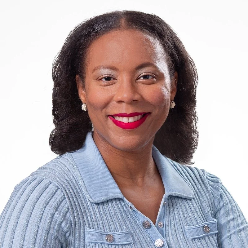 A professional woman with dark curly hair, wearing a light blue button-up shirt and earrings, smiling against a plain white background.