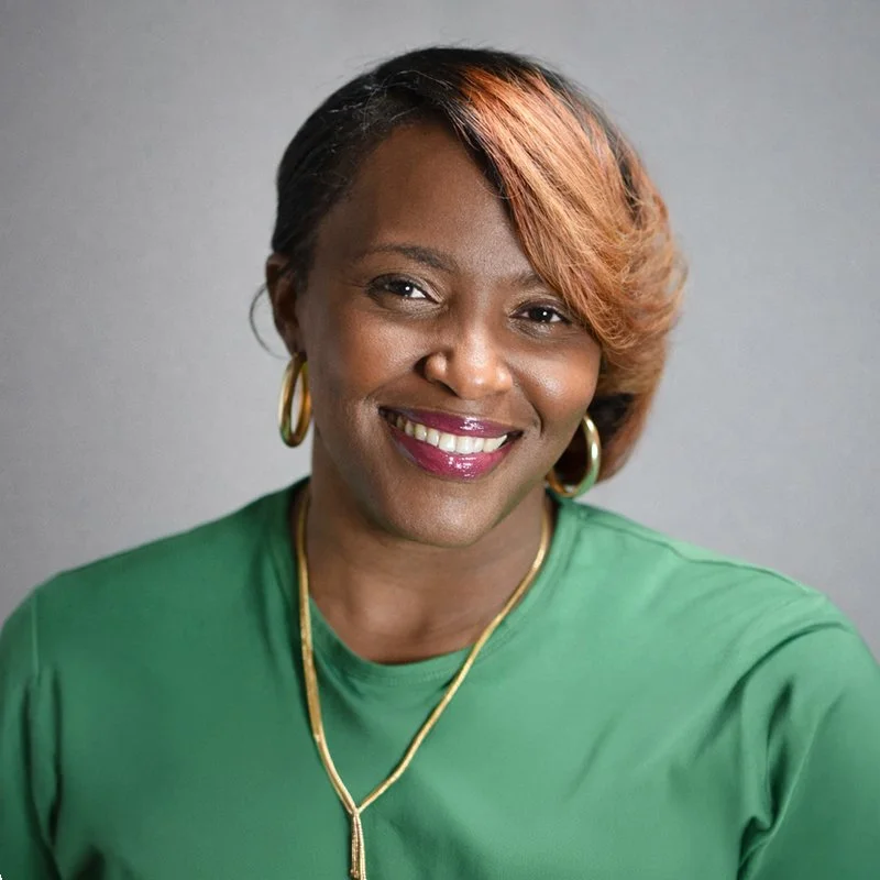 A woman with short, styled hair with highlights, smiling and wearing a green shirt, gold hoop earrings, and a gold necklace, standing against a plain gray background.