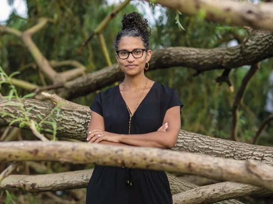 Woman with glasses, dark curly hair in bun, wearing black dress, standing outdoors among fallen tree branches and green foliage.