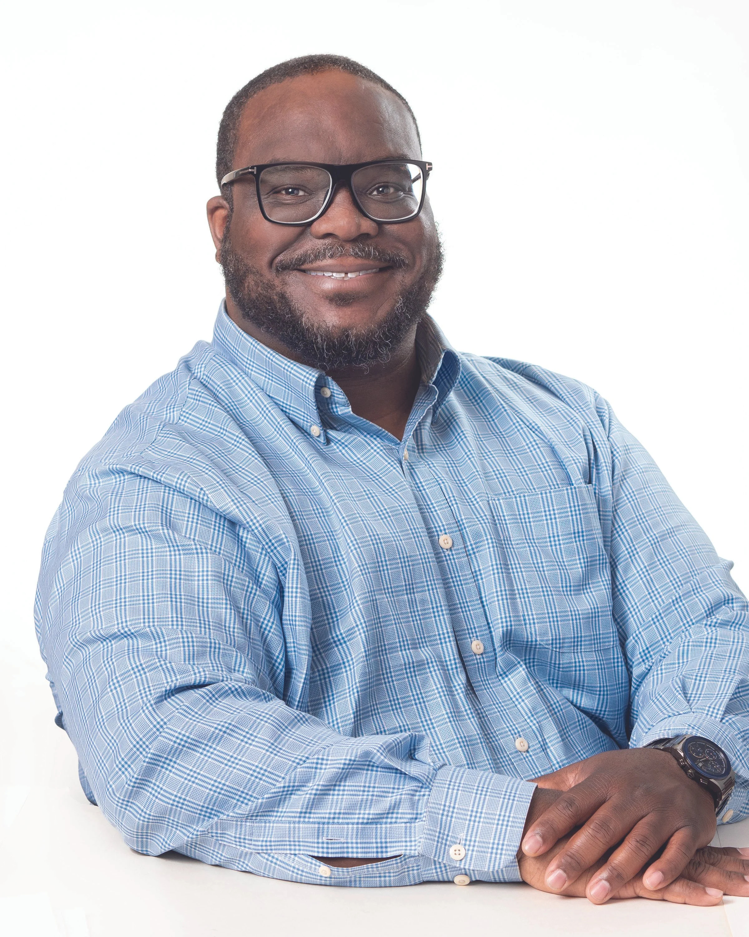 A man wearing glasses and a blue checkered shirt smiling with his hands clasped on a white table against a plain white background.
