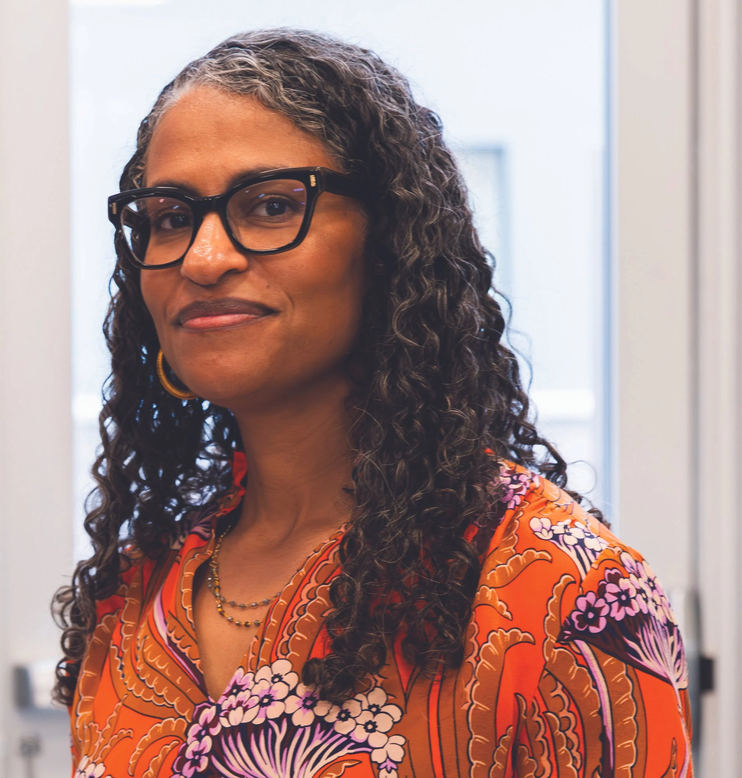 A woman with curly hair and glasses wearing a colorful floral-patterned shirt, standing indoors near a window.