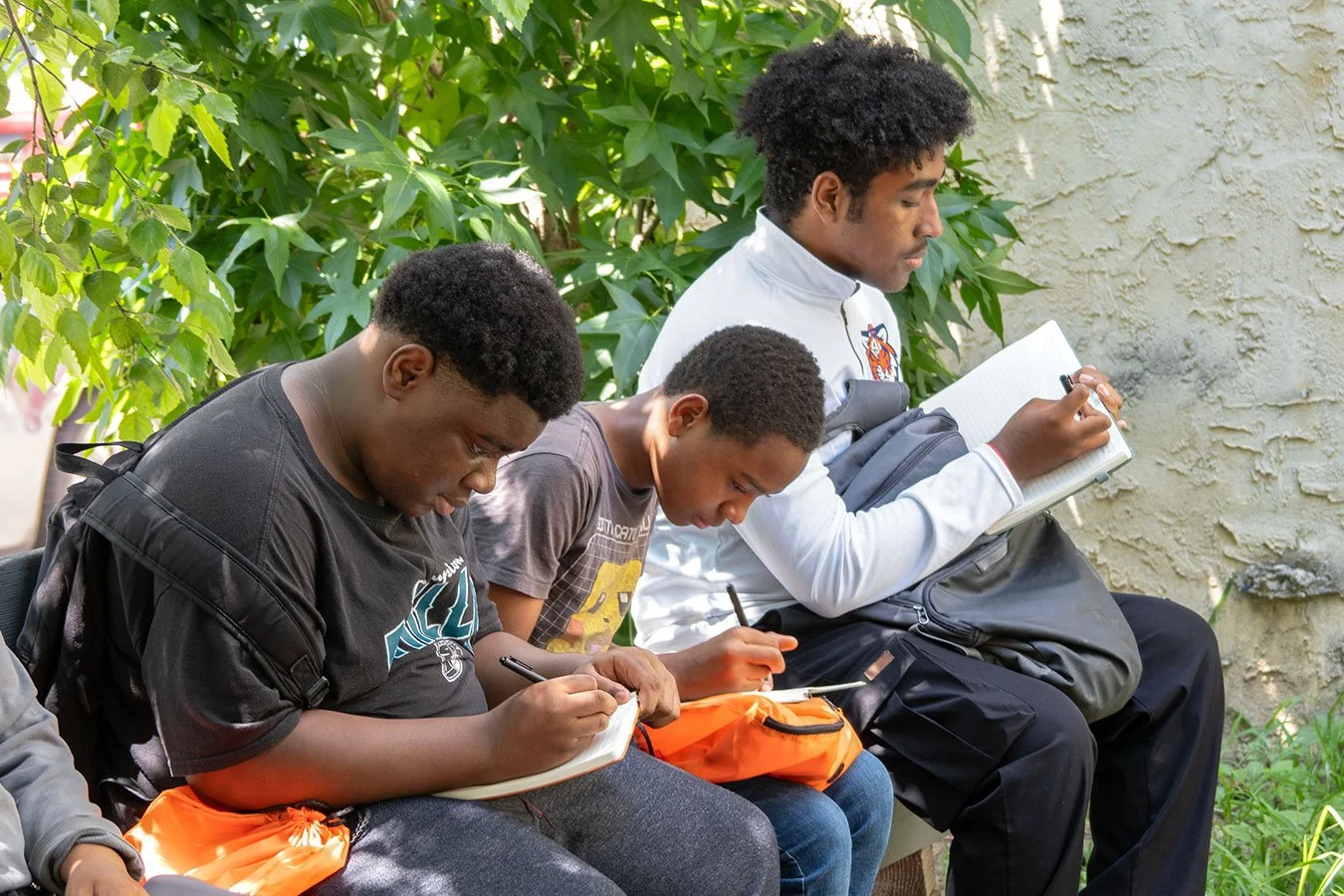 Three young males sitting on a bench outdoors under green leafy trees, writing in notebooks with backpacks nearby.